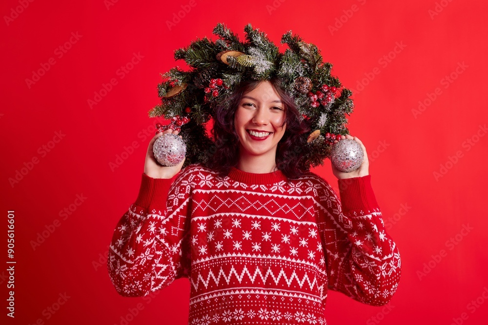 Expressive young woman with dark hair wearing an ugly Christmas sweater and a wreath headband. She is holding silver ornaments in her hands and smiling at the camera. Isolated on red background. Expressive young woman with dark hair wearing an ugly Christmas sweater and a wreath headband. She is holding silver ornaments in her hands and smiling at the camera. Isolated on red background.