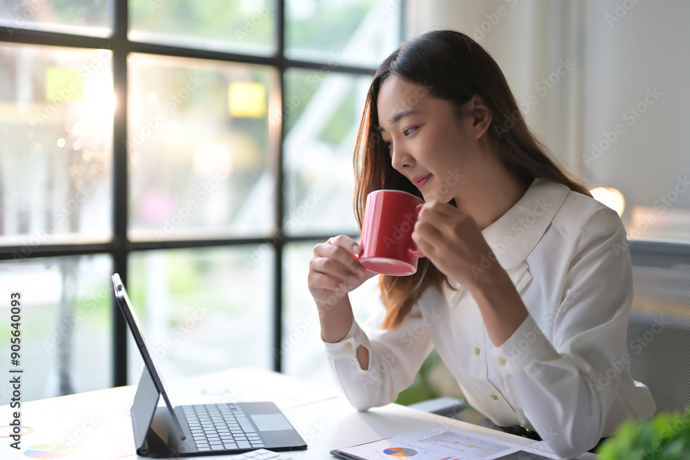 Young Professional Woman Working on Laptop and Drinking Coffee in Modern Office with Natural Light Young Professional Woman Working on Laptop and Drinking Coffee in Modern Office with Natural Light