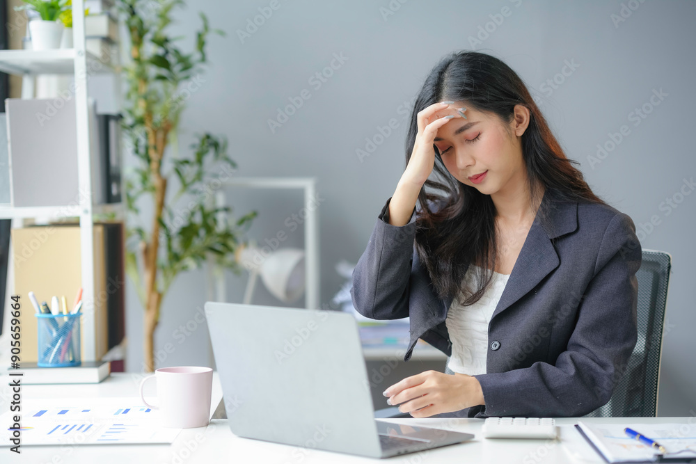 Young businesswoman is feeling tired and having a headache while she is working on a laptop at her desk in the office Young businesswoman is feeling tired and having a headache while she is working on a laptop at her desk in the office