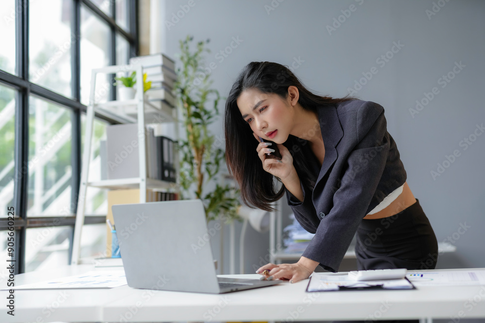 Young businesswoman is multitasking in her modern office, talking on the phone while working on her laptop Young businesswoman is multitasking in her modern office, talking on the phone while working on her laptop
