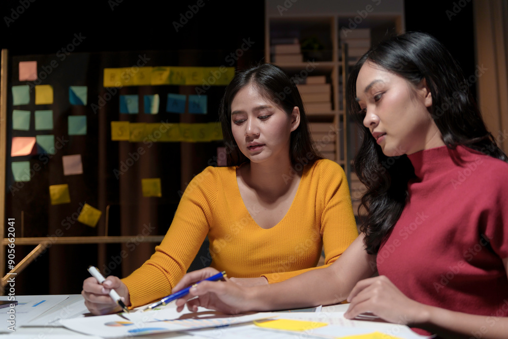 Two young businesswomen reviewing data and brainstorming in their office at night Two young businesswomen reviewing data and brainstorming in their office at night