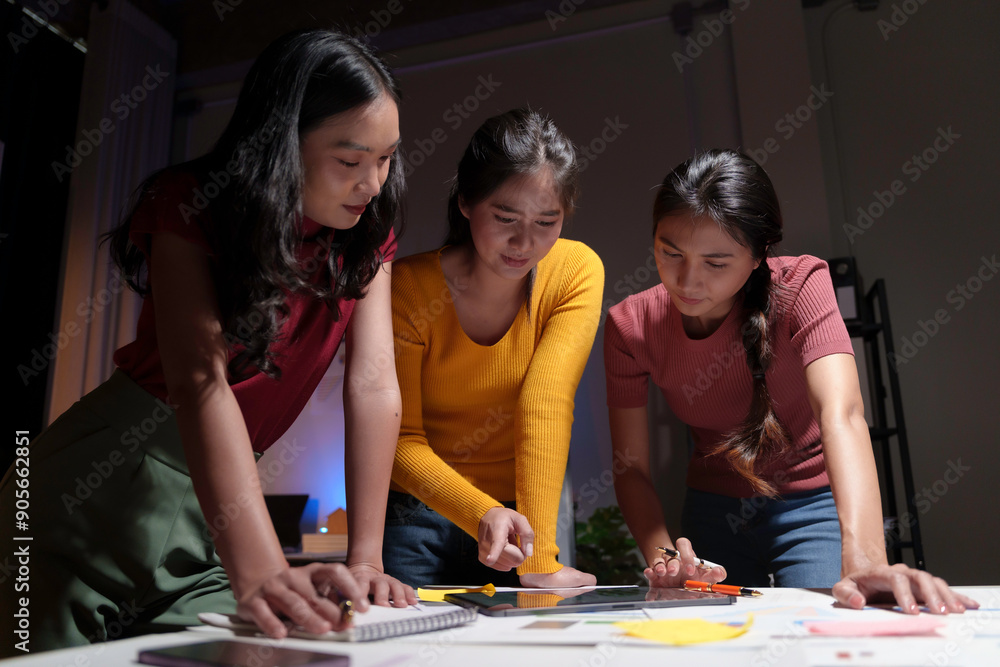 Three asian businesswomen leaning over a table and reviewing paperwork while working late in an office at night Three asian businesswomen leaning over a table and reviewing paperwork while working late in an office at night