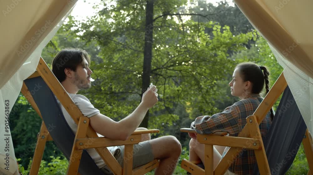 Young couple is seen vaping in nature, sitting in chairs in front of their tent. Medium shot. Slow motion. Young couple is seen vaping in nature, sitting in chairs in front of their tent. Medium shot. Slow motion.