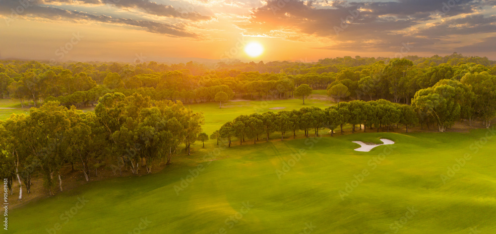 Obraz premium Panorama of golf course at sunset with dramatic sky. Scenic panoramic view of golf fairway with pond. Golf field with beautiful pines