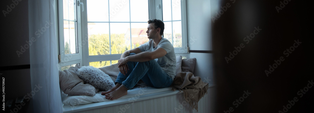 Fototapeta premium Depressed, sad, frustrated, crying, scared, instability mental and bipolar young man sitting on the windowsill at home. Headache because overworked and stress