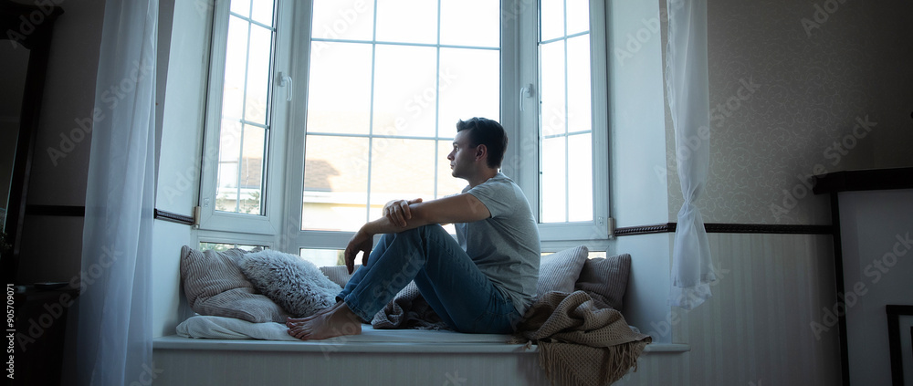 Depressed, sad, frustrated, crying, scared, instability mental and bipolar young man sitting on the windowsill at home. Headache because overworked and stress Depressed, sad, frustrated, crying, scared, instability mental and bipolar young man sitting on the windowsill at home. Headache because overworked and stress