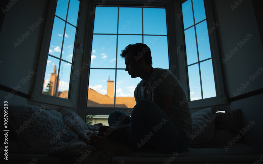Silhouette side view image of depressed, sad, frustrated, crying, scared, instability mental and bipolar young man sitting on the windowsill at home. Headache because overworked and stress Silhouette side view image of depressed, sad, frustrated, crying, scared, instability mental and bipolar young man sitting on the windowsill at home. Headache because overworked and stress