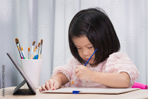 A little girl is happily drawing with paintbrush at a table, surrounded by colorful art supplies. Asian child aged 3-4 years old. Homeschooling. Kid learning art. photo