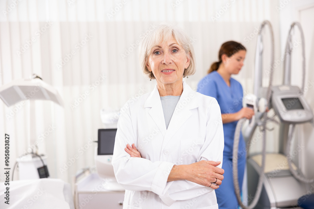 Obraz premium Smiling old woman doctor posing against background of cosmetology devices in aesthetic clinic