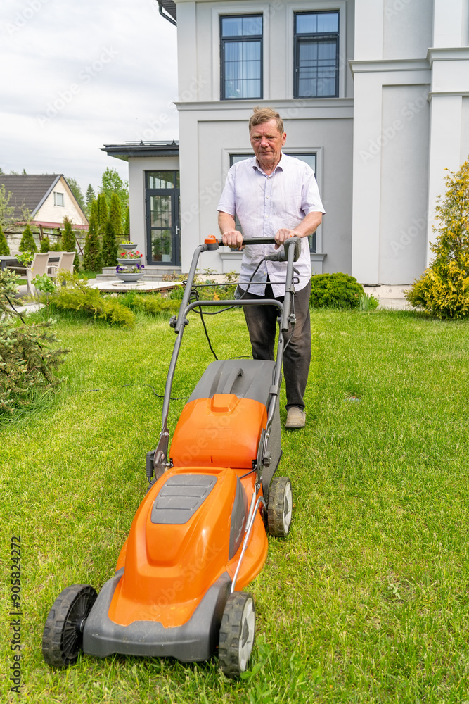 Fototapeta premium An elderly man trims his vibrant lawn with a mower beside his contemporary home