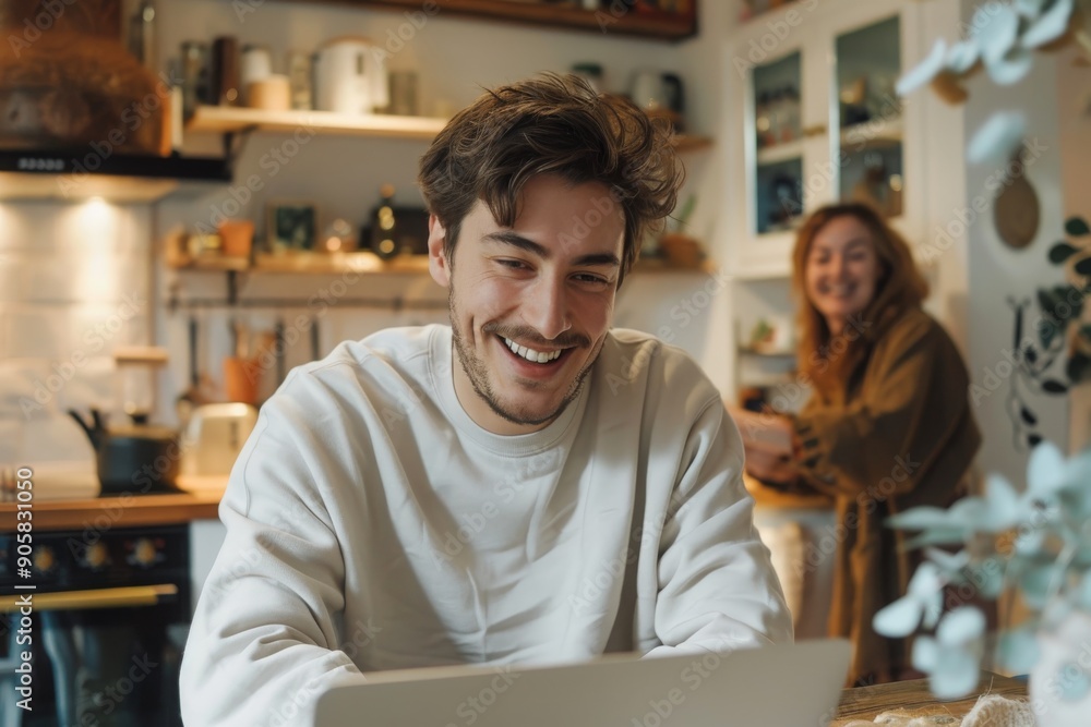 Young Man Laughing While Using Laptop in Kitchen Young Man Laughing While Using Laptop in Kitchen