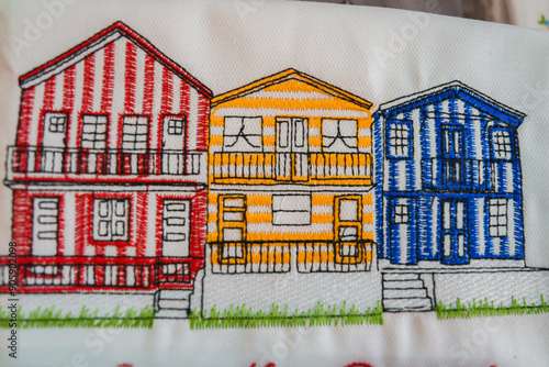 Detailed embroidery of three vibrant Costa Nova beach houses in Aveiro, Portugal. Features red, yellow, and blue vertical stripes, balconies, and multiple windows.