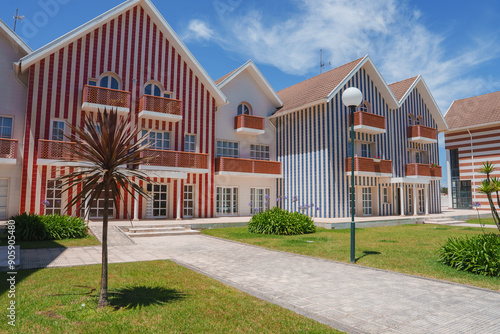 A row of distinctive palheiros with red and blue vertical stripes in Aveiro, Portugal. The scene features a bright blue sky, scattered clouds, and a well-maintained lawn. photo