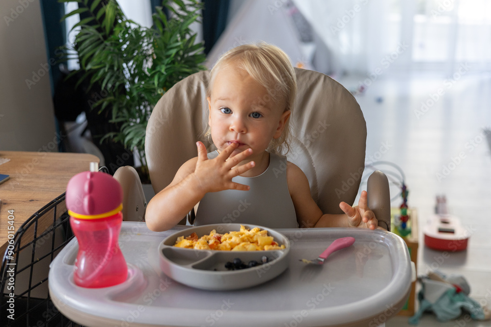 a blonde baby eats with a spoon while sitting at a children's dining table a blonde baby eats with a spoon while sitting at a children's dining table