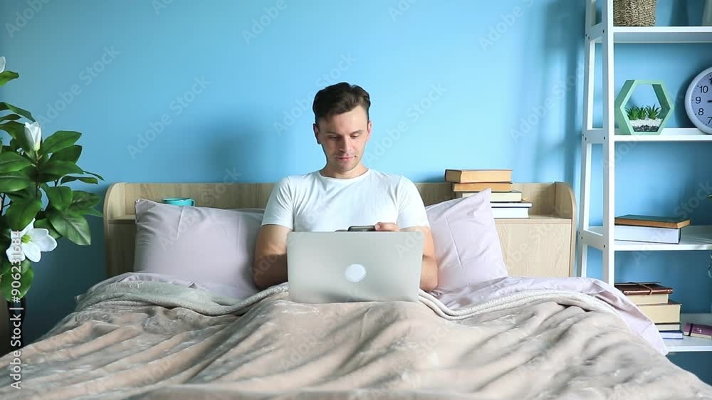 Young modern handsome man working on laptop in the bed early in the morning. Bet, online shopping, freelancer, watch the movie, studying, office at home Young modern handsome man working on laptop in the bed early in the morning. Bet, online shopping, freelancer, watch the movie, studying, office at home