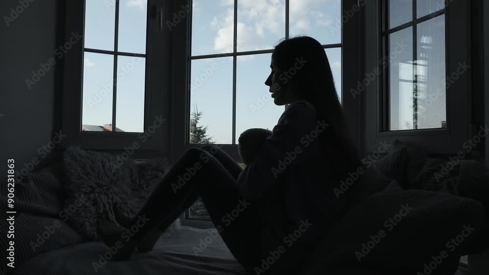 Shadowed silhouette of a depressed woman while she sitting on windowsill with wide window on background. Sad, crisis, bad emotions, mental health, family violence. Shadowed silhouette of a depressed woman while she sitting on windowsill with wide window on background. Sad, crisis, bad emotions, mental health, family violence.