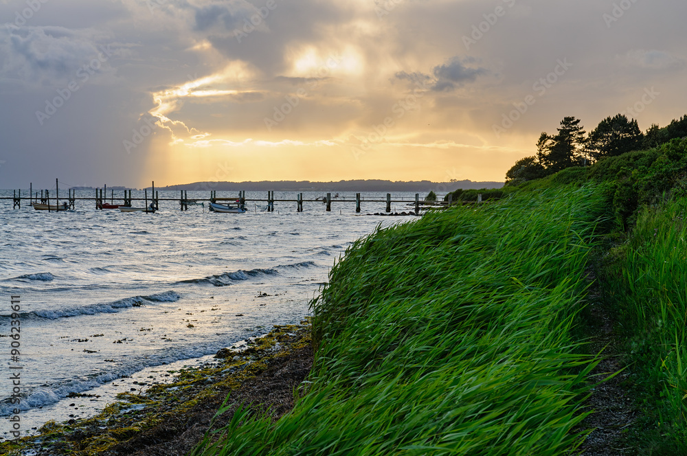 Gentle waves lap at the shore as sunlight filters through dark clouds, casting a warm glow over the peaceful waterfront dock and lush greenery. Gentle waves lap at the shore as sunlight filters through dark clouds, casting a warm glow over the peaceful waterfront dock and lush greenery.