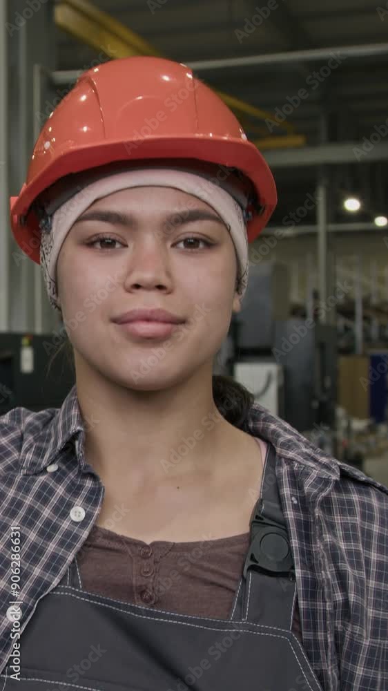 Vertical slowmo portrait of young Latin workwoman in safety overalls looking confidently at camera, fixing orange hard hat on head in manufacturing factory Vertical slowmo portrait of young Latin workwoman in safety overalls looking confidently at camera, fixing orange hard hat on head in manufacturing factory
