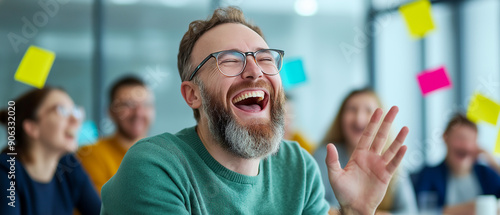 Group of young professionals laughing together during a meeting in a modern office. The man in focus has a big smile, wearing glasses and a green sweater, creating a lively and positive atmosphere.