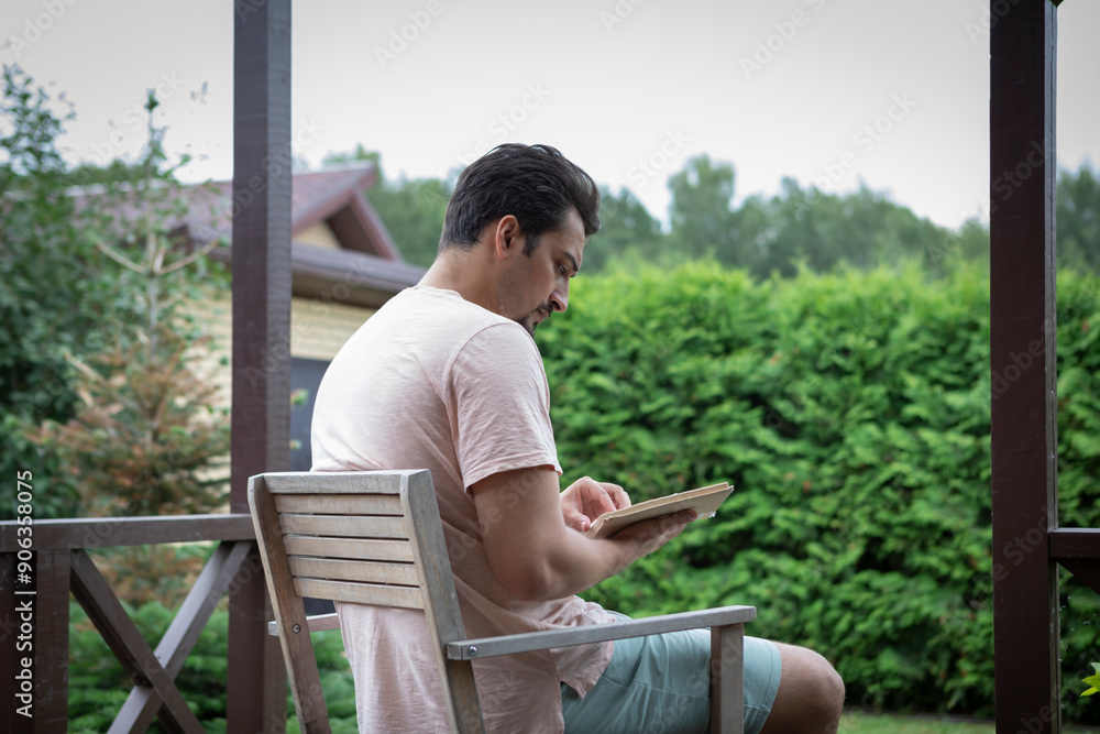 A young man sits on the veranda of a country house in good weather and reads an interesting book A young man sits on the veranda of a country house in good weather and reads an interesting book