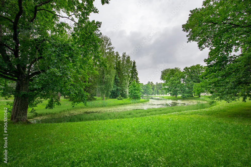 A tranquil park features vibrant green grass and towering trees, while a gentle waterway flows through the lush landscape under overcast skies. A tranquil park features vibrant green grass and towering trees, while a gentle waterway flows through the lush landscape under overcast skies.