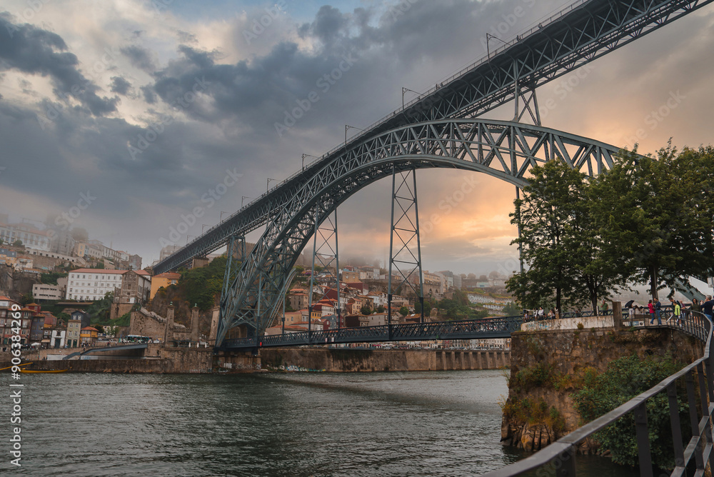 Naklejka premium The iconic Dom Luis I Bridge spans the Douro River in Porto, Portugal, with a warm sunset glow. Traditional hillside buildings and a serene riverbank enhance the scene.