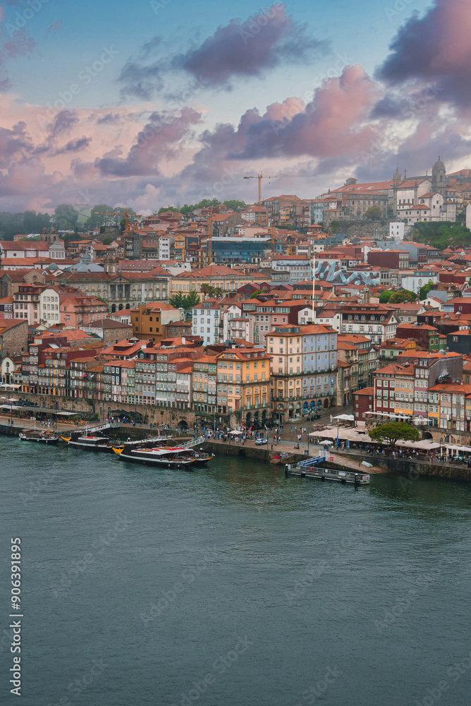 Fototapeta premium Colorful historic buildings with red-tiled roofs line the bustling waterfront of Porto's Ribeira district, featuring boats, a church spire, and dramatic clouds.