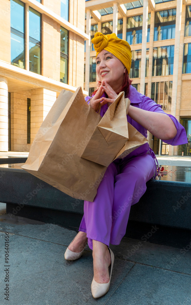 woman with craft shopping bags in the city. The concept of fashionable Consumerism and ecology. An adult Asian woman in a purple suit with red and yellow accents enjoys shopping . a sunny day.