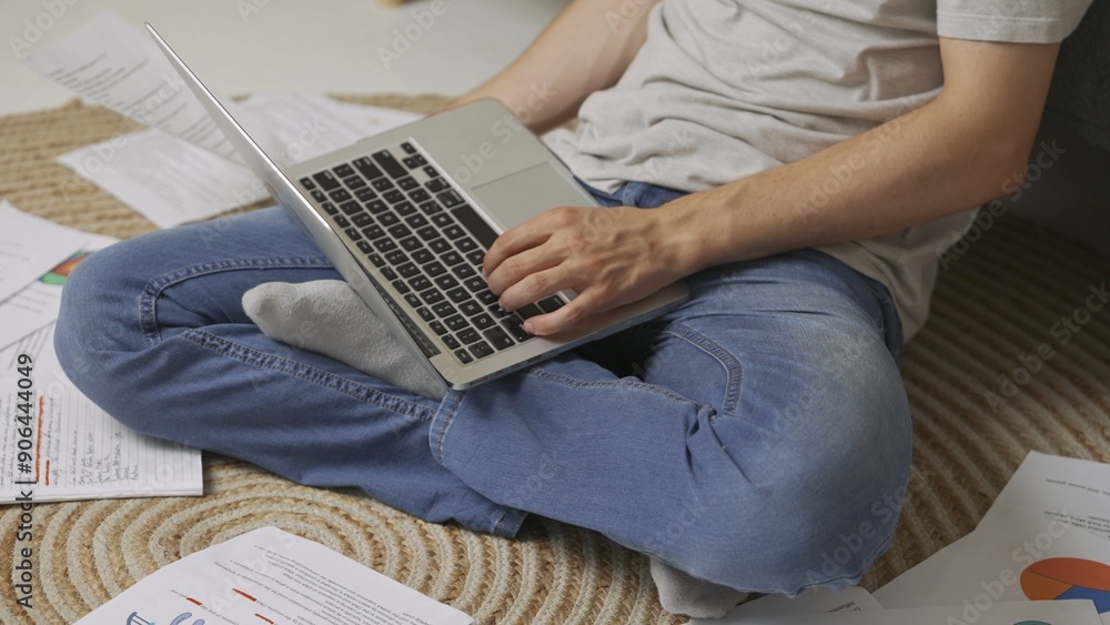 Naklejka premium Focused adult man college student sitting on the floor in apartment room at daylight and doing homework typing on laptop. Education concept.