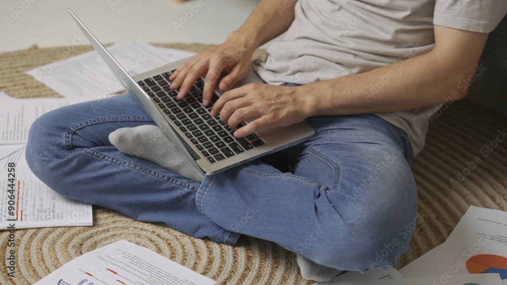 Naklejka premium Focused adult man college student sitting on the floor in apartment room at daylight and doing homework typing on laptop. Education concept.