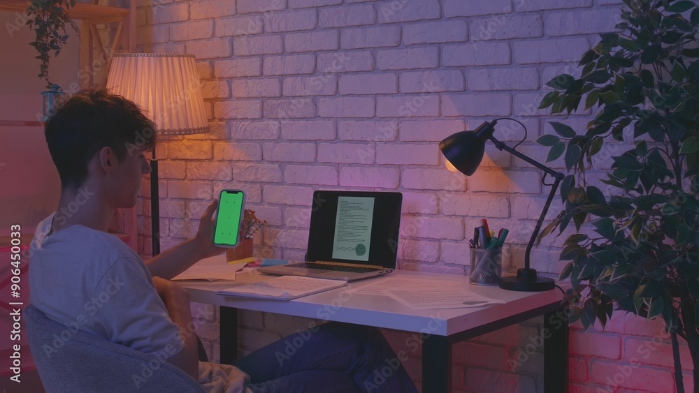 Fototapeta premium Focused adult man college student sitting at desk in apartment room late at night and doing homework using smartphone chroma key green screen mockup. Education concept.