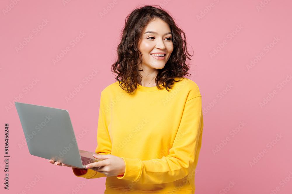 Naklejka premium Young fun happy IT woman she wear yellow sweatshirt casual clothes hold use work on laptop pc computer look aside on area isolated on pastel light pink background studio portrait. Lifestyle concept.