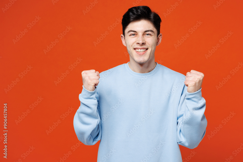 Young smiling happy Caucasian man he wear blue sweatshirt casual clothes doing winner gesture celebrate clenching fists say yes isolated on plain red orange color background studio. Lifestyle concept. Young smiling happy Caucasian man he wear blue sweatshirt casual clothes doing winner gesture celebrate clenching fists say yes isolated on plain red orange color background studio. Lifestyle concept.