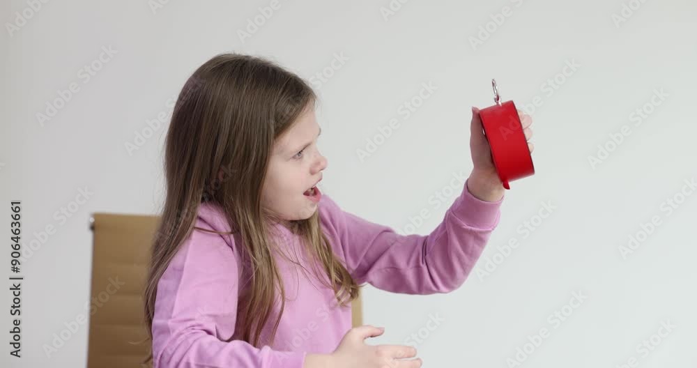 Curious little girl shakes alarm clock sitting at wooden desk in room slow motion. Child tries to stop ringing expressing irritation Curious little girl shakes alarm clock sitting at wooden desk in room slow motion. Child tries to stop ringing expressing irritation