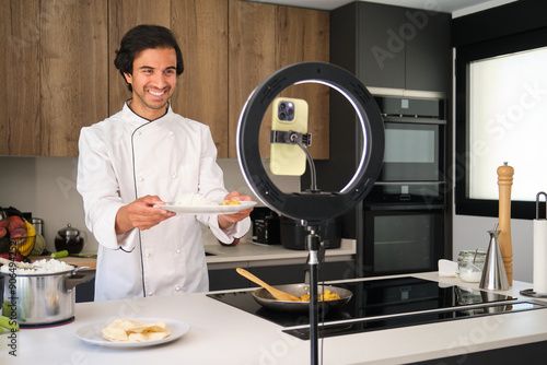 Obraz na płótnie Indian chef is holding a plate and smiling while recording a culinary show in a modern kitchen w salonie