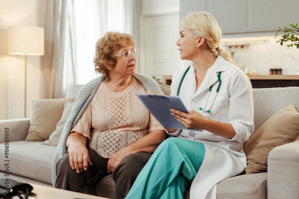Obraz premium Female doctor wearing lab coat holding medical chart speaking with senior patient during home visit
