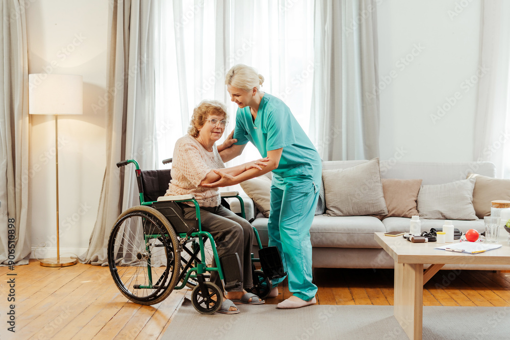 Smiling nurse assisting senior woman getting up from wheelchair at home Smiling nurse assisting senior woman getting up from wheelchair at home