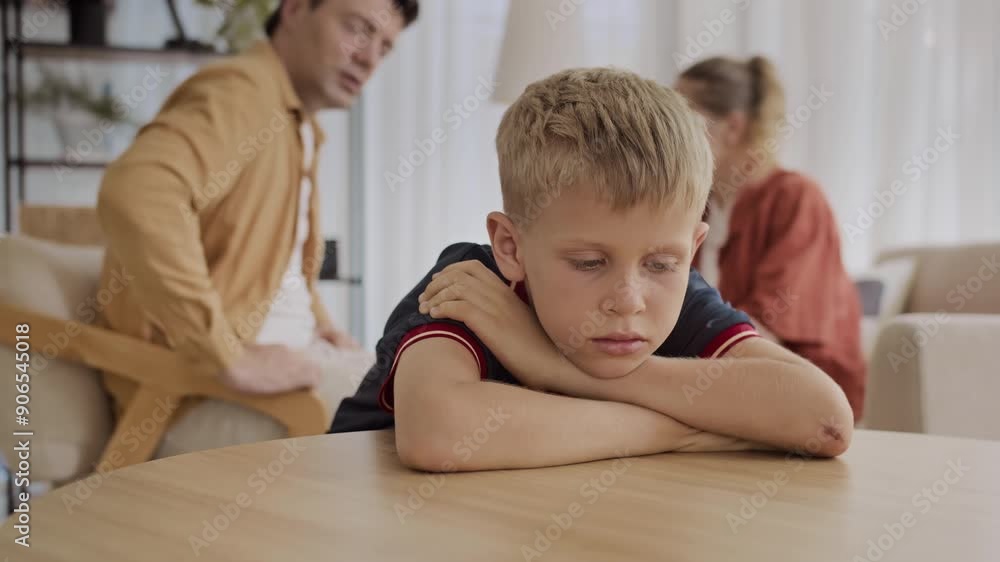 Medium shot of frustrated Caucasian teenage kid sitting at table while his parents having quarrel in background at home Medium shot of frustrated Caucasian teenage kid sitting at table while his parents having quarrel in background at home