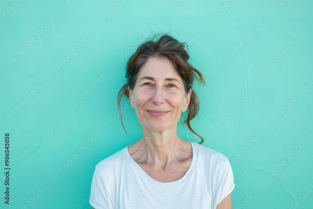 A cheerful middle-aged woman stands against a vibrant turquoise wall, exuding warmth and positivity in a simple white T-shirt. A cheerful middle-aged woman stands against a vibrant turquoise wall, exuding warmth and positivity in a simple white T-shirt.