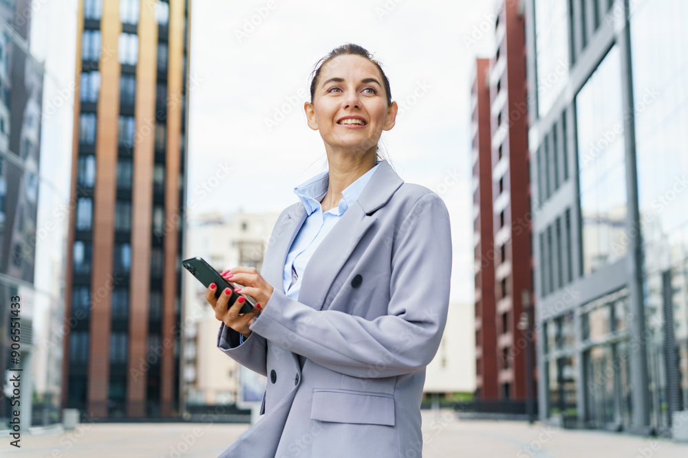 Fototapeta premium Confident businesswoman with a smartphone, standing between modern office buildings, exuding professionalism and success.