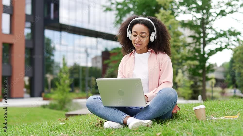 African american female student works on laptop sitting on lawn in campus space near university building. Young black woman in headphones learns remotely, listens to distance seminar, lecture, lesson African american female student works on laptop sitting on lawn in campus space near university building. Young black woman in headphones learns remotely, listens to distance seminar, lecture, lesson