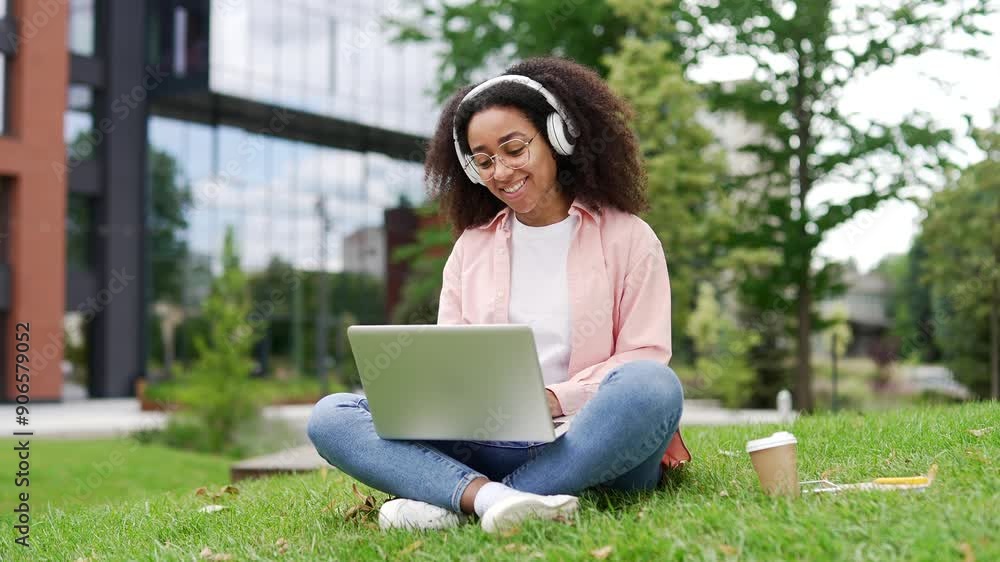 African american female student talking on video call using a laptop sitting on the lawn in campus near university building. Happy young black woman in headphones greeting, smiling, chatting online African american female student talking on video call using a laptop sitting on the lawn in campus near university building. Happy young black woman in headphones greeting, smiling, chatting online