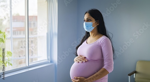 Pregnant Indian woman wearing a face mask standing in a clinic room with hand on her belly and gazing out the window, healthcare setting, expectant mother