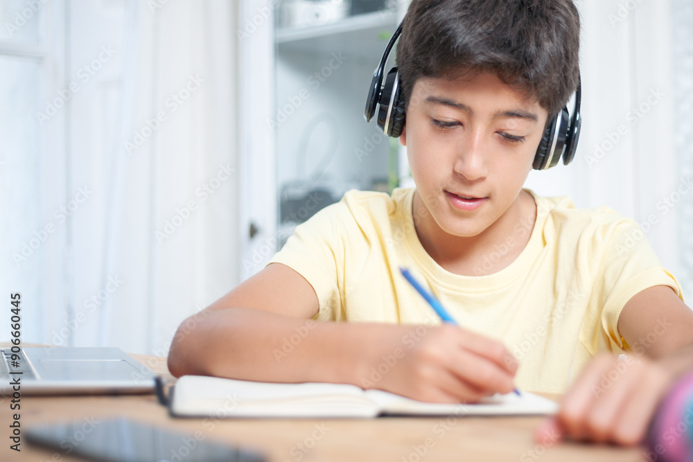 Naklejka premium Young student writing with a pen on a notepad on a desk. She is wearing a wireless headset.