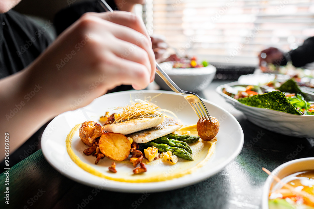A close-up of hands using utensils to eat a gourmet dish with fish, asparagus, and sliced potatoes on a white plate. The meal is well-presented, healthy, and suitable for food-related content. A close-up of hands using utensils to eat a gourmet dish with fish, asparagus, and sliced potatoes on a white plate. The meal is well-presented, healthy, and suitable for food-related content.