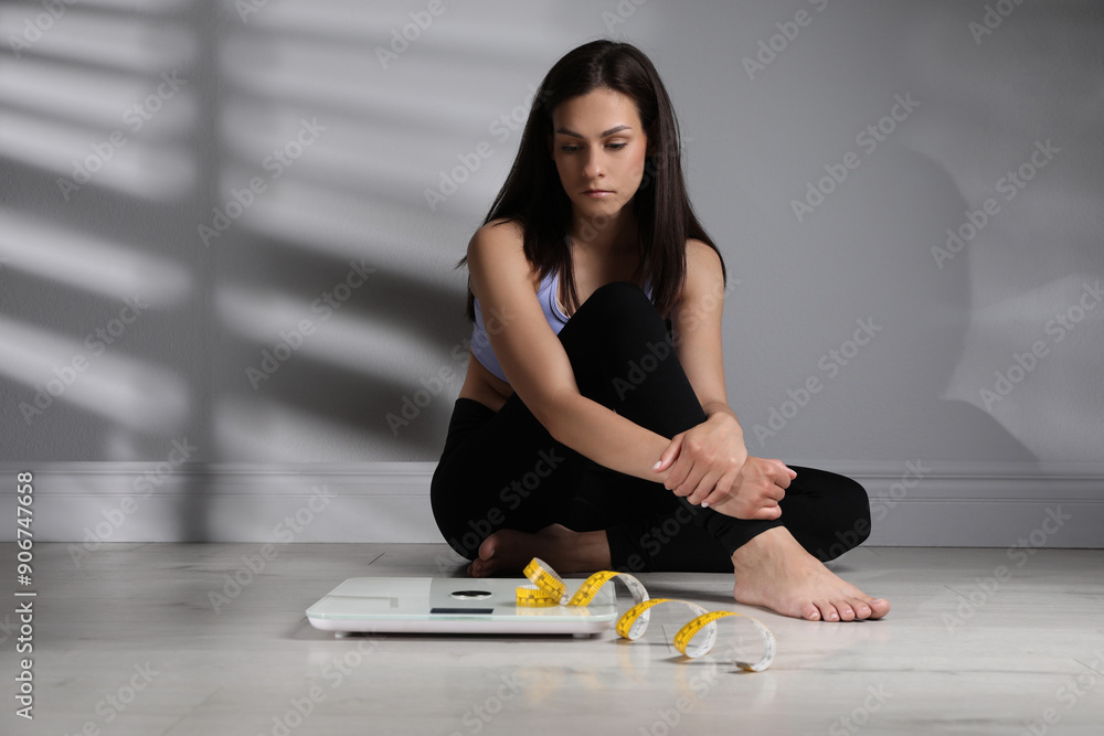 Eating disorder. Sad woman sitting near scale and measuring tape on floor indoors Eating disorder. Sad woman sitting near scale and measuring tape on floor indoors