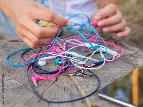 A phone and a hand holding a knot of four pairs of in-ear wired colorful pink, blue, white and black earbuds tangled in a messy chaotic problem. photo