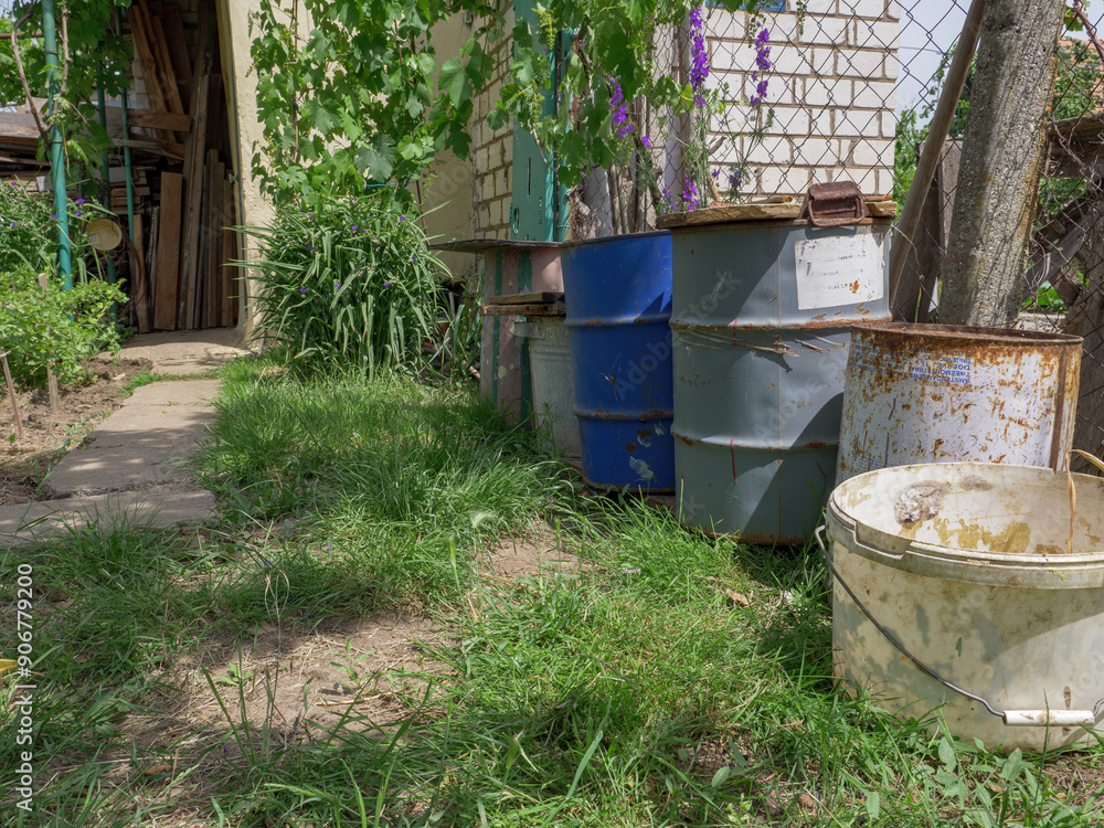 Old rusty metal barrels with wooden lids and plastic buckets as rural storage at the back yard of a country house. Old rusty metal barrels with wooden lids and plastic buckets as rural storage at the back yard of a country house.