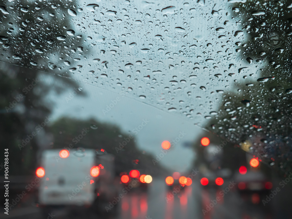 Closeup of rain drops on a windshield, and blurry road traffic with cars and red and orange lights. View from the driver's seat.