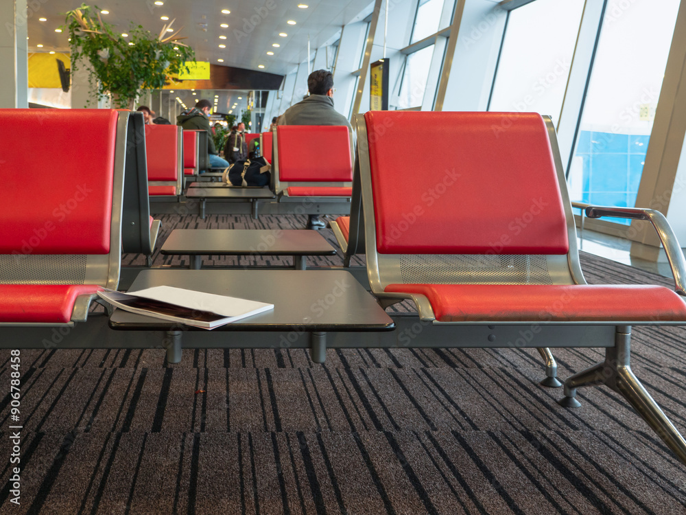 Rows of red seats at an airport terminal. Magazine lying at a table between chairs. People sitting and waiting planes boarding.